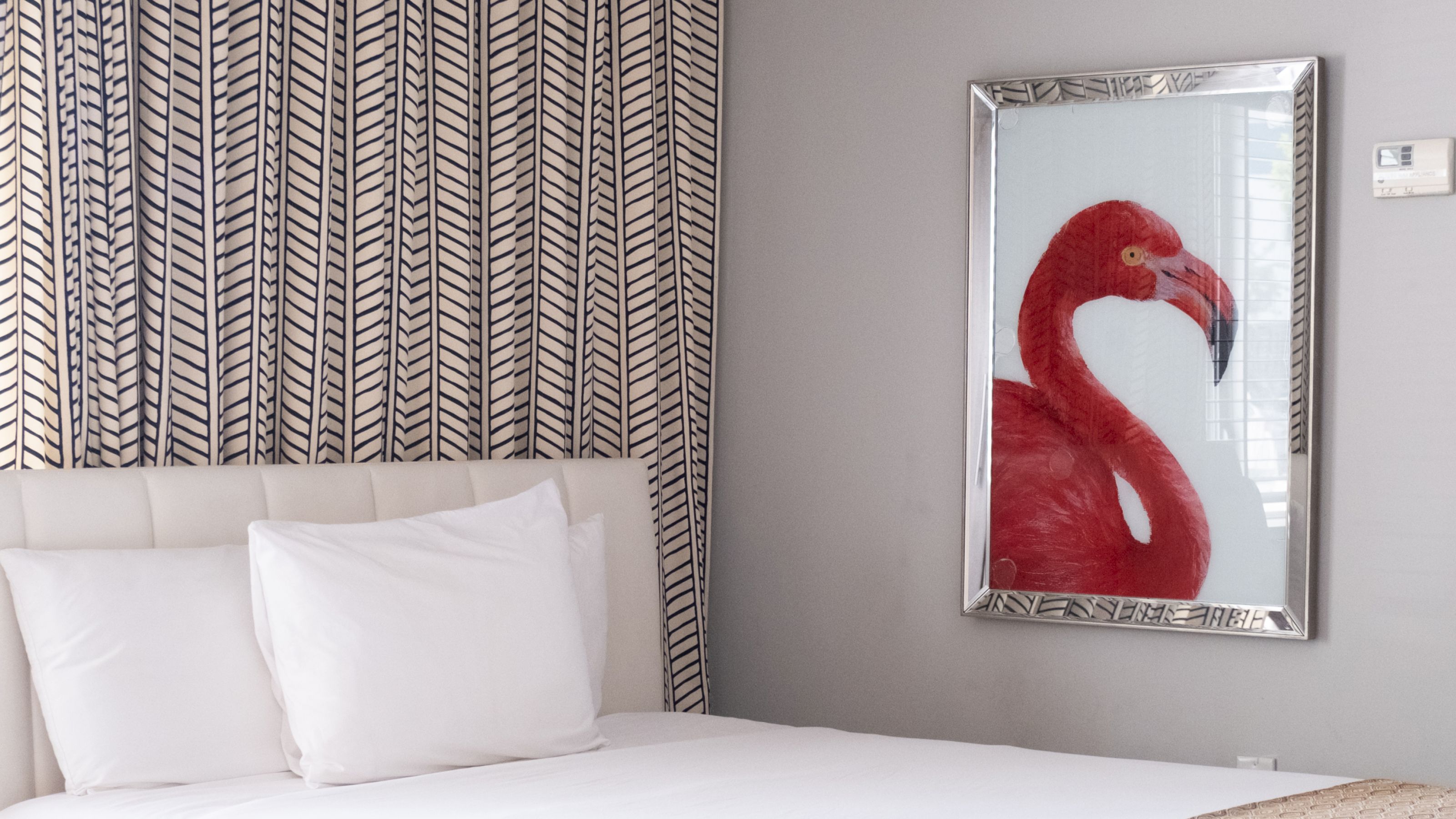 A neatly made bed with white linens and pillows, a patterned headboard, and a framed flamingo artwork on the wall.