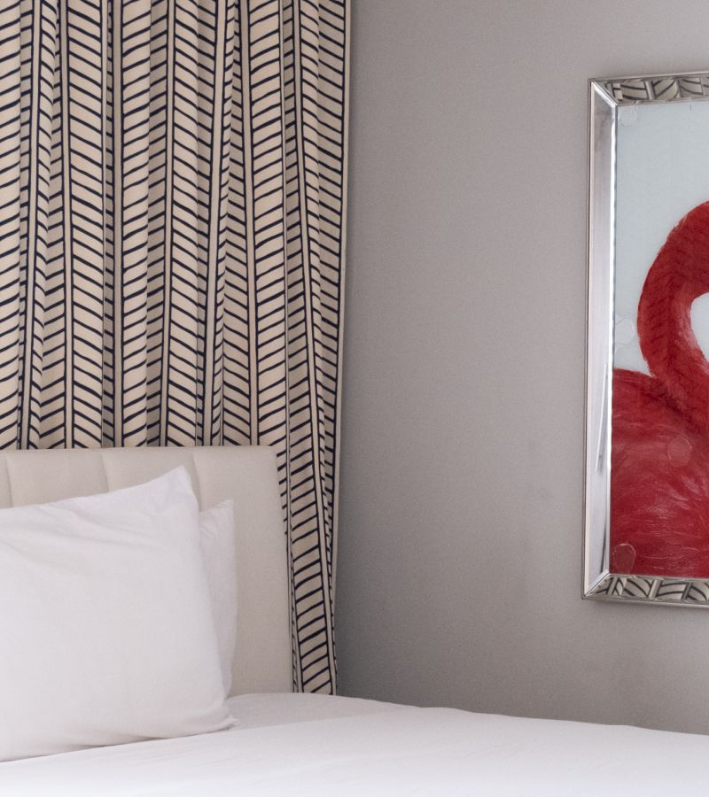 A neatly made bed with a white duvet and pillow, a patterned curtain, and a framed flamingo artwork on the wall.