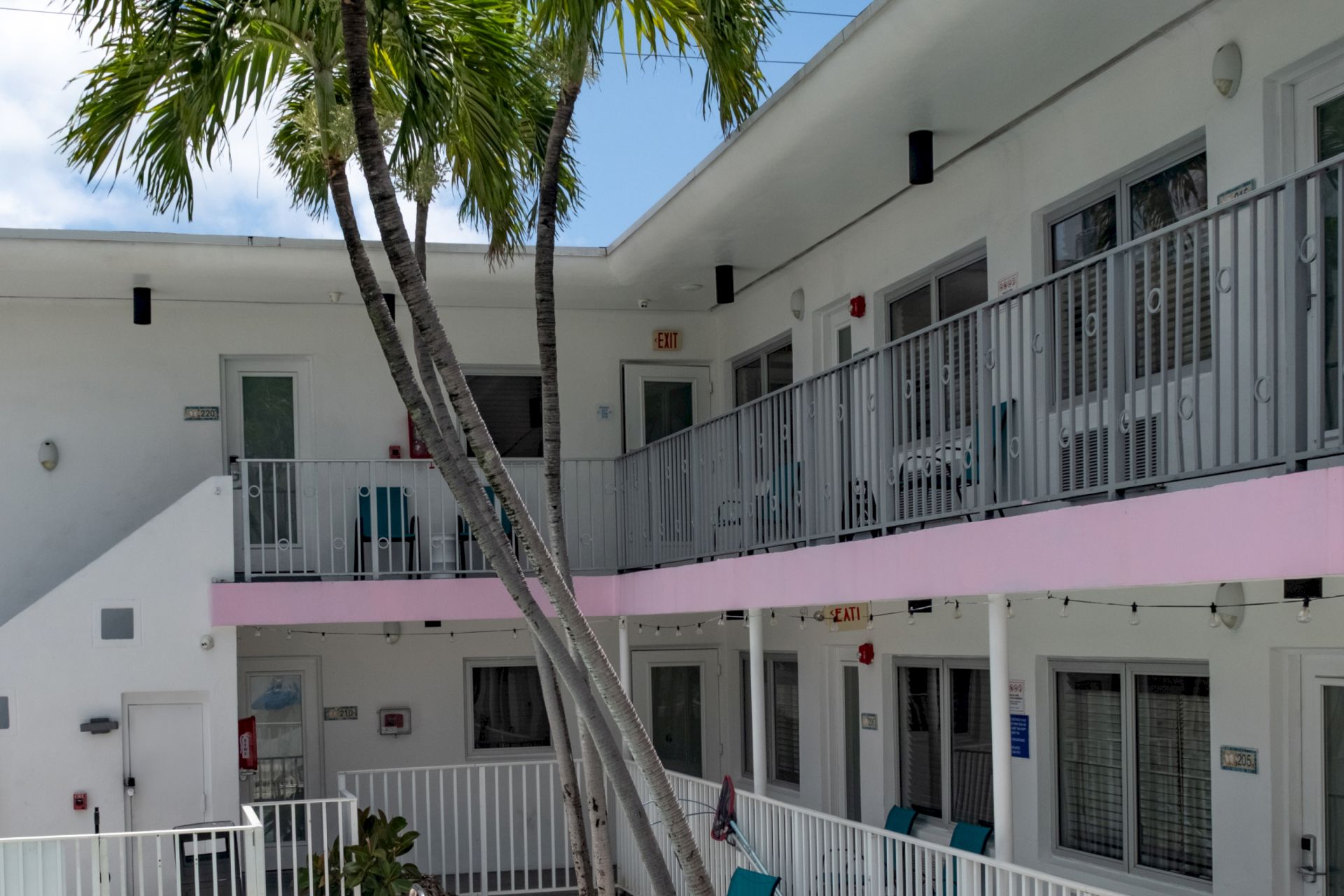 A white building with pink accents and a balcony overlooking a courtyard with palm trees.