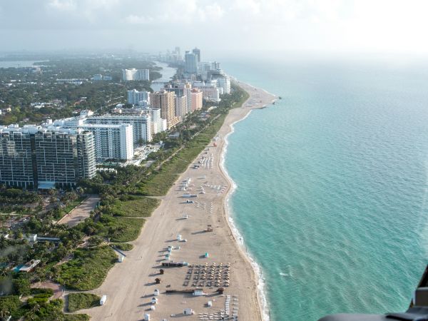 Aerial view of a beach with rows of umbrellas and high-rise buildings along the coastline under a blue sky with clouds.