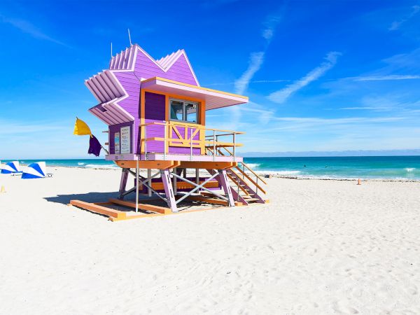 A vibrant purple lifeguard tower with yellow accents on a sandy beach, under a clear blue sky with a yellow and purple flag flying.