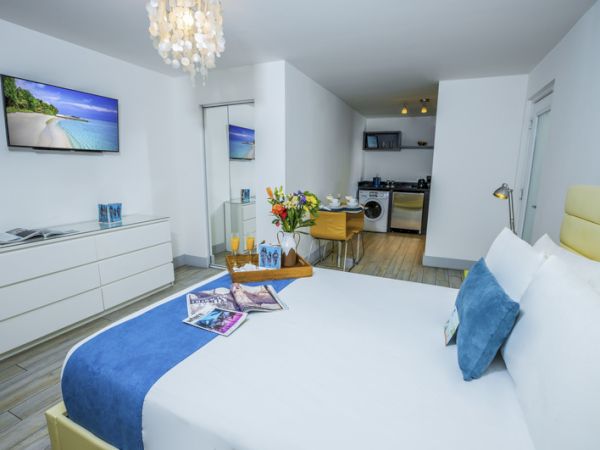 The image shows a modern bedroom with a large bed, flat-screen TV, dresser, dining table, and a kitchenette in the background.