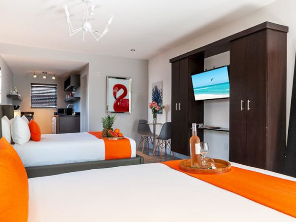 The image shows a modern hotel room with two beds, bright orange accents, a wall-mounted TV, a dining area, and a small kitchenette.