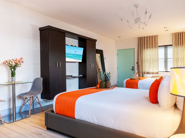 A modern hotel room with two beds, orange accents, a TV, flowers, and a small table with chairs completes the setup.