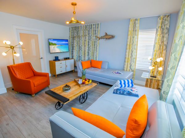 A modern living room with orange and gray furniture, a wall-mounted TV, decorative items on a wooden coffee table, and yellow curtains.