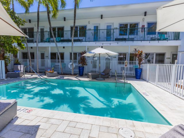 A rectangular pool with lounge chairs and umbrellas in front of a white building with balconies.