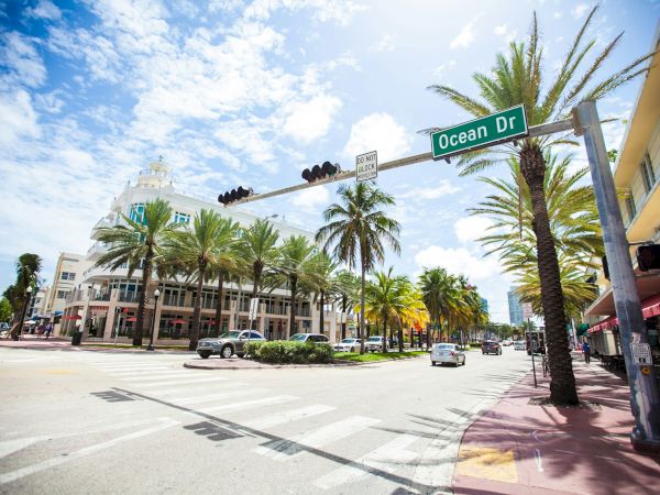 A sunny street scene with palm trees, cars, a pedestrian crossing, and an "Ocean Dr" sign under a partly cloudy sky.