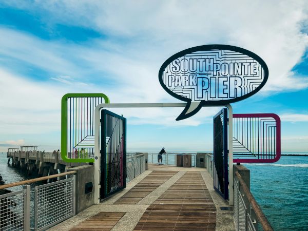 A pier entrance with the sign "South Pointe Park Pier" in a speech bubble, located over ocean water and surrounded by colorful frames.
