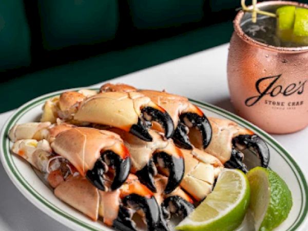 A plate of stone crab claws with lime wedges next to a copper cup labeled "Joe's," containing a drink garnished with lime.