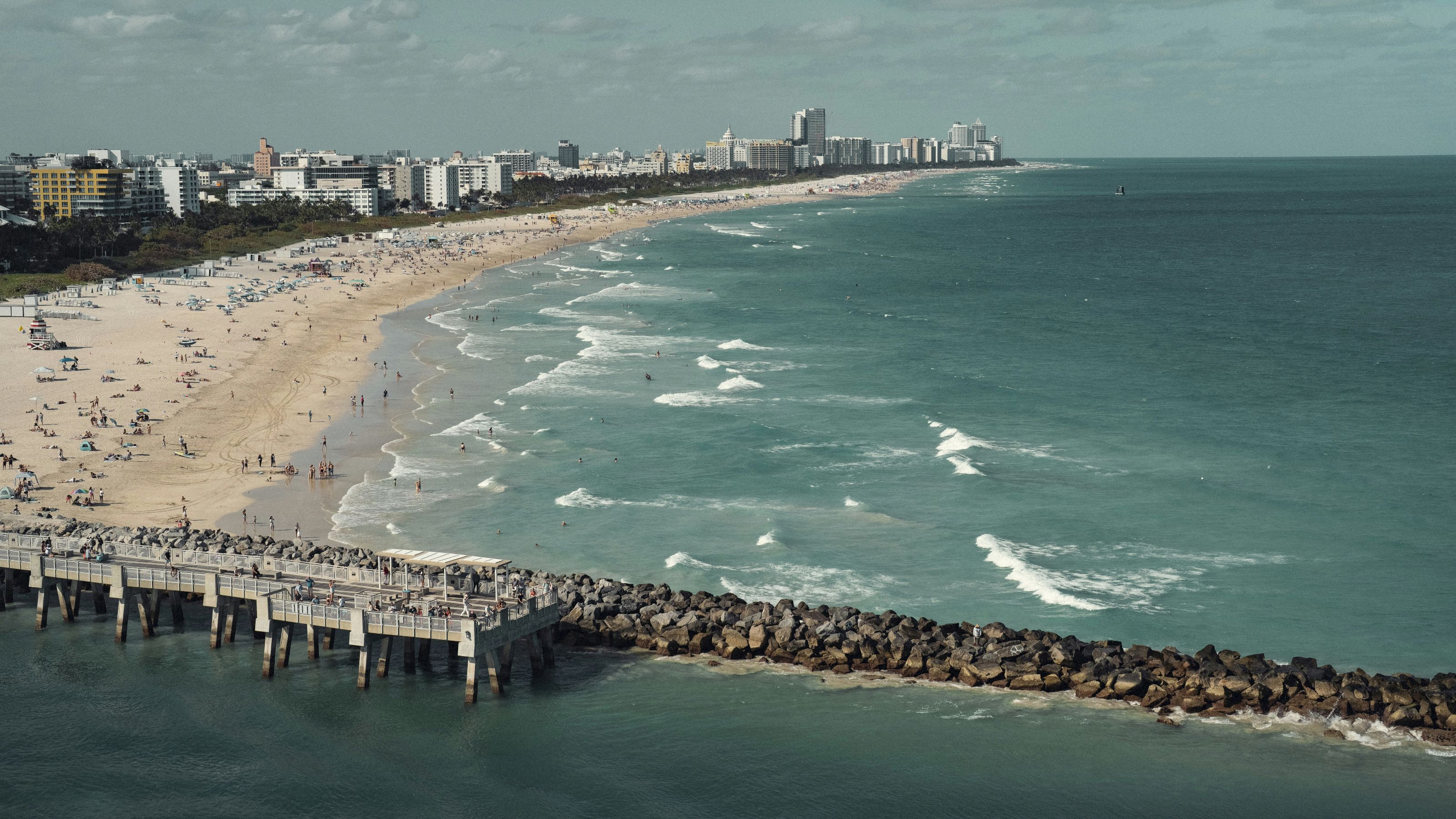 A wide aerial view of a beach with a pier and city skyline in the background.