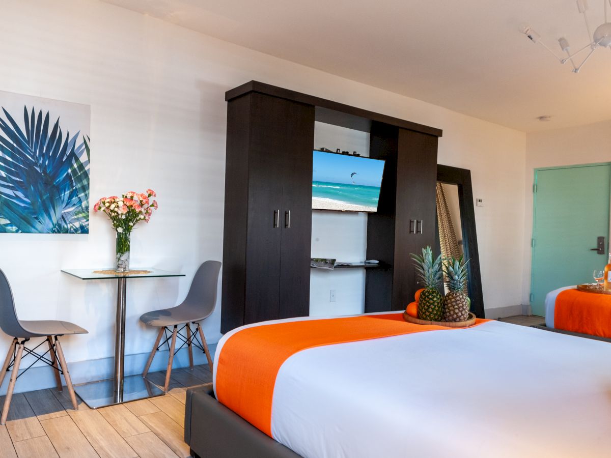 A bright, modern hotel room with two neatly made beds featuring white and orange bedding, a dark wood wardrobe unit with a TV, a small table with chairs and decorative plants, and a mint-green door in the background.
