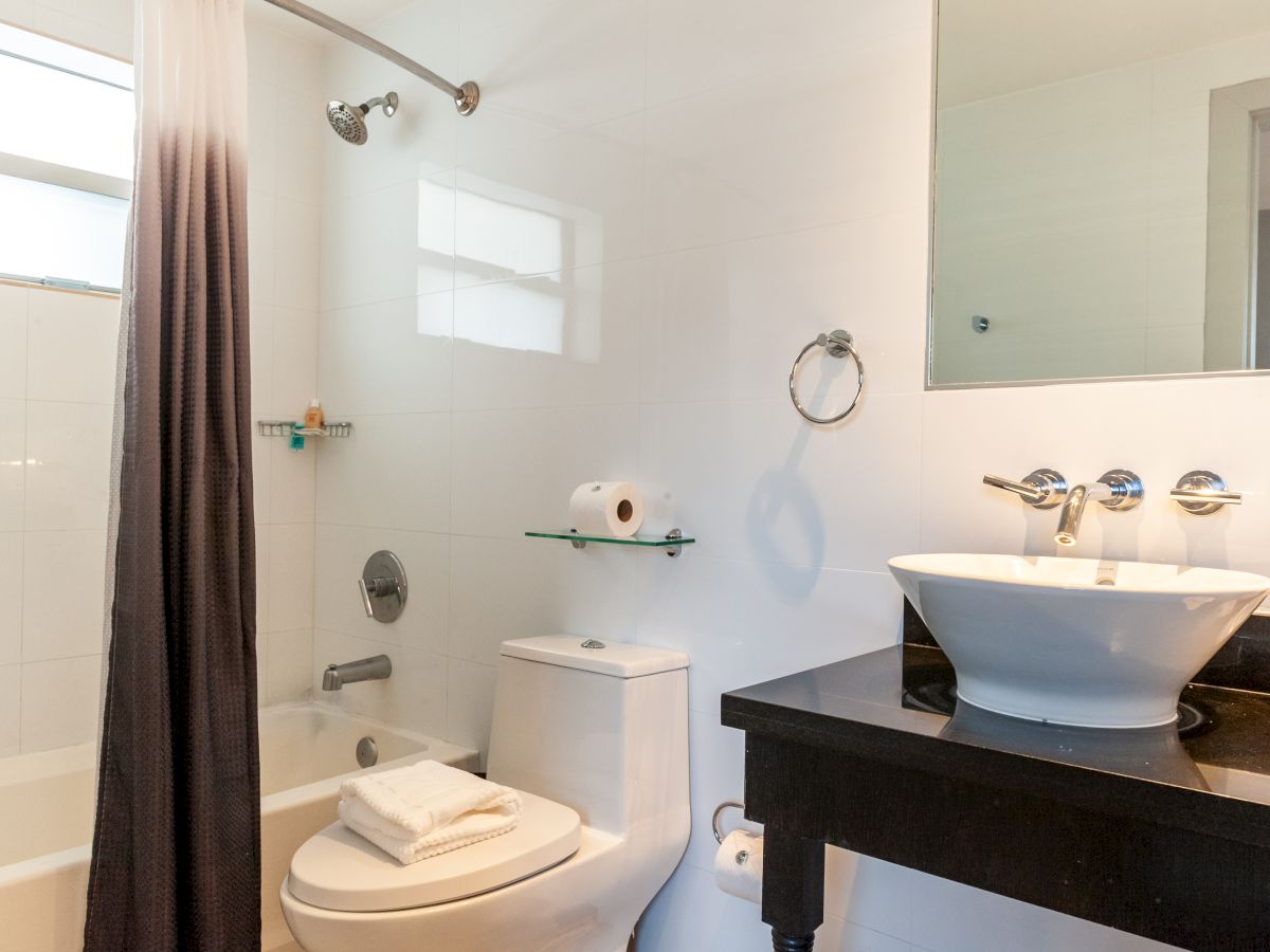 A modern bathroom with a bathtub/shower, white tiles, a toilet, a black vanity with a vessel sink, a round mirror, and towels neatly stacked on the counter.