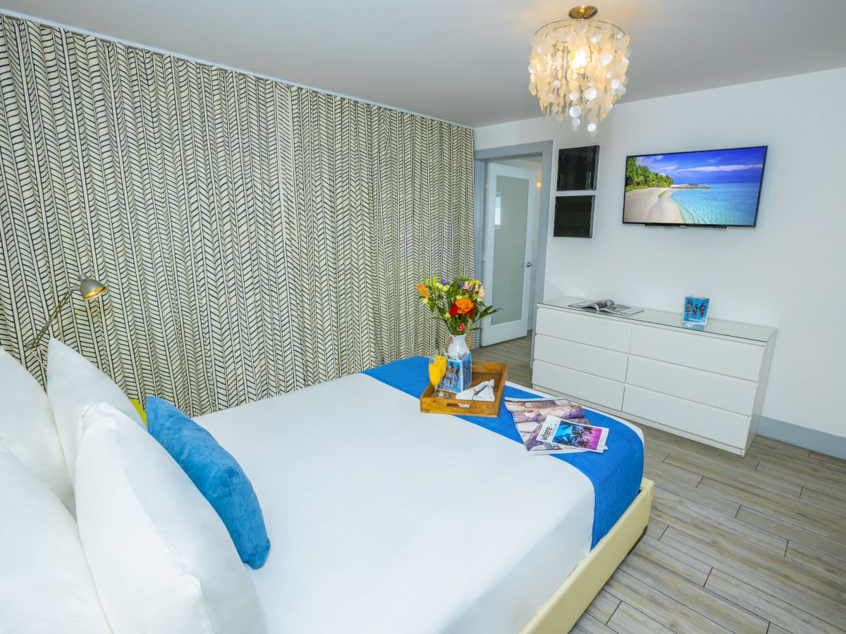 A bright hotel room with a double bed, white bedding and blue accents, a dresser with a TV, flowers on a table, and a chandelier, modern and tidy.