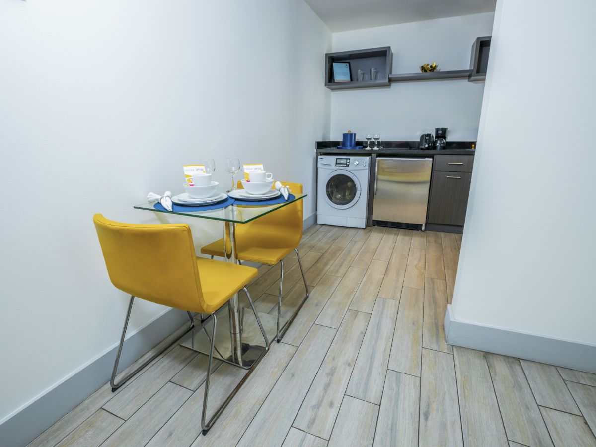 A compact kitchen-dining area with a small glass table set for two, yellow chairs, white walls, a washer and dishwasher, and a counter with shelves above.