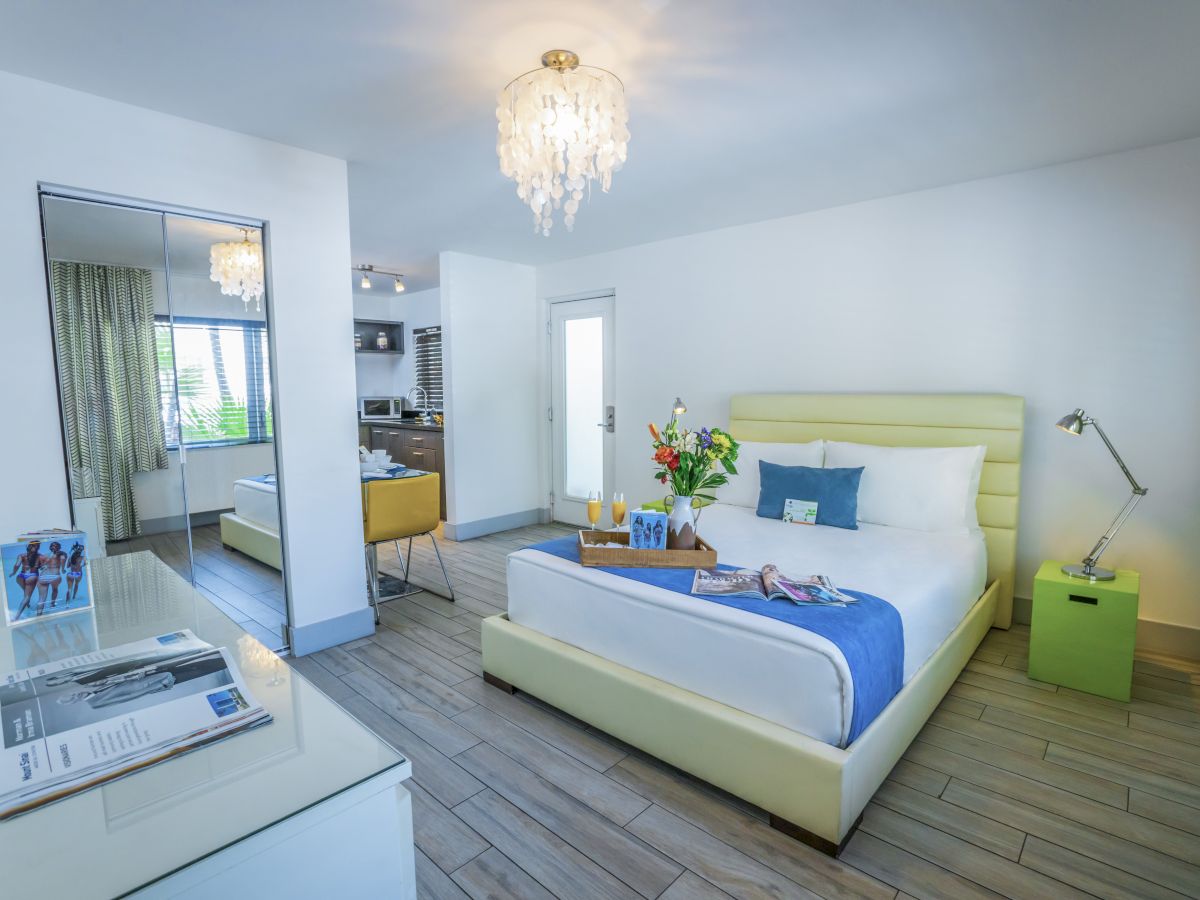 A bright modern hotel room with a pale yellow bed, blue and white bedding, a chandelier, a desk, and a small lime nightstand, airy and stylish.