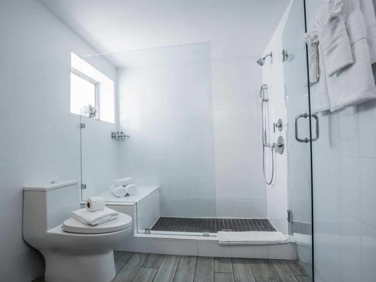 A modern, bright bathroom with a glass shower enclosure, a toilet, a small vanity, towels, and minimalist white tiles, all neatly arranged.