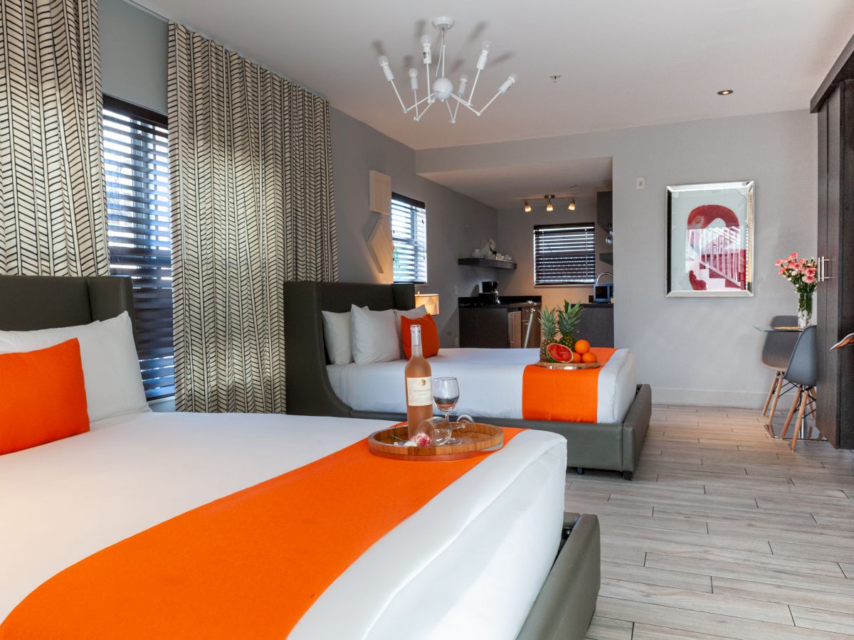 A bright, stylish hotel room with a king bed and orange accents, a seating area with a grey sofa, contemporary art, and a minibar in a modern, airy space.