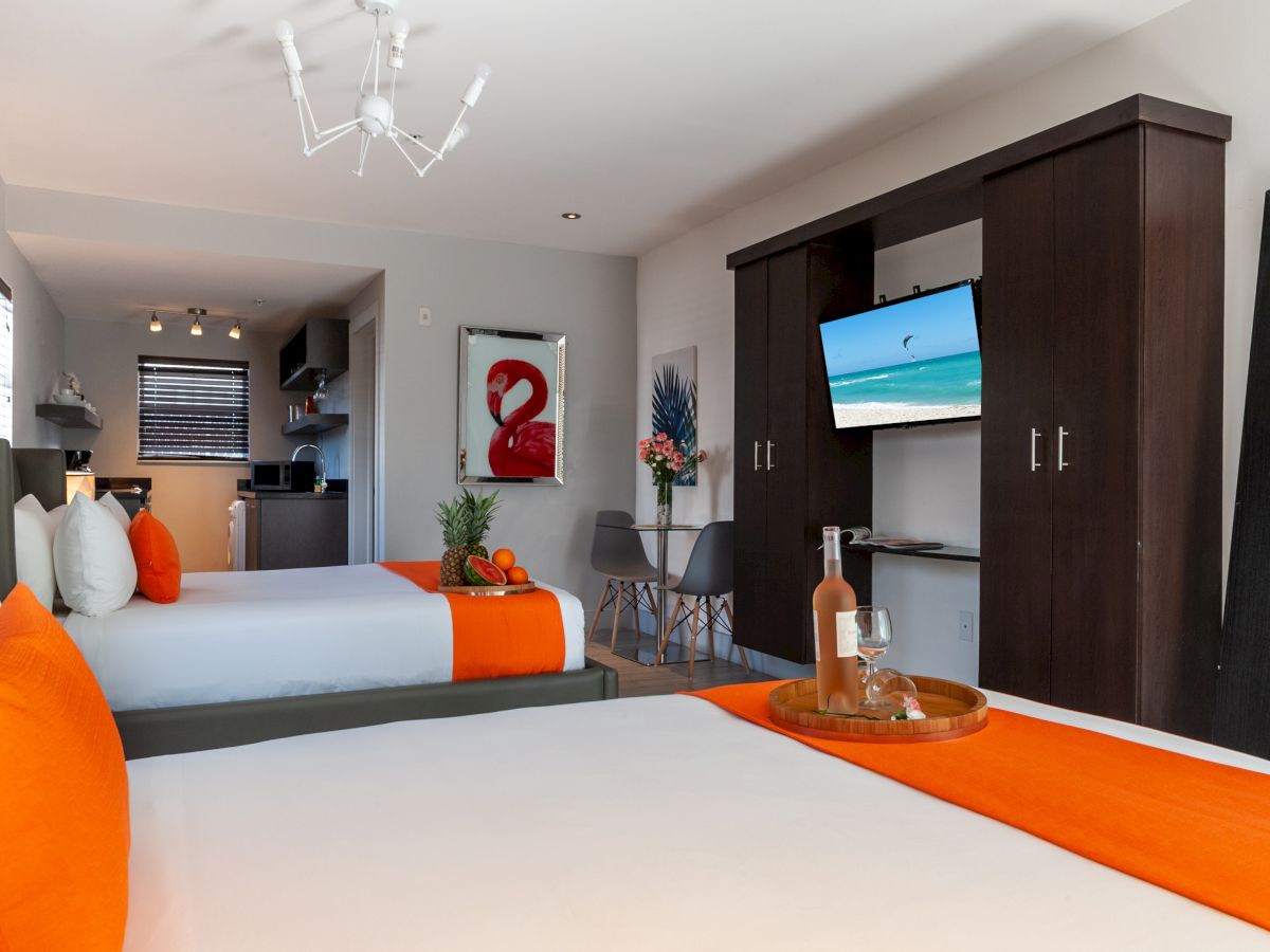 A modern hotel room with two beds, orange accents, a TV mounted in a dark wood cabinet, and a small seating area in the back.