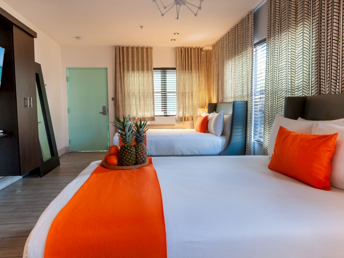 Top it at 140 characters, ending with a period: A bright hotel room with two beds, bold orange accents, pineapple centerpiece, and large windows with patterned curtains.