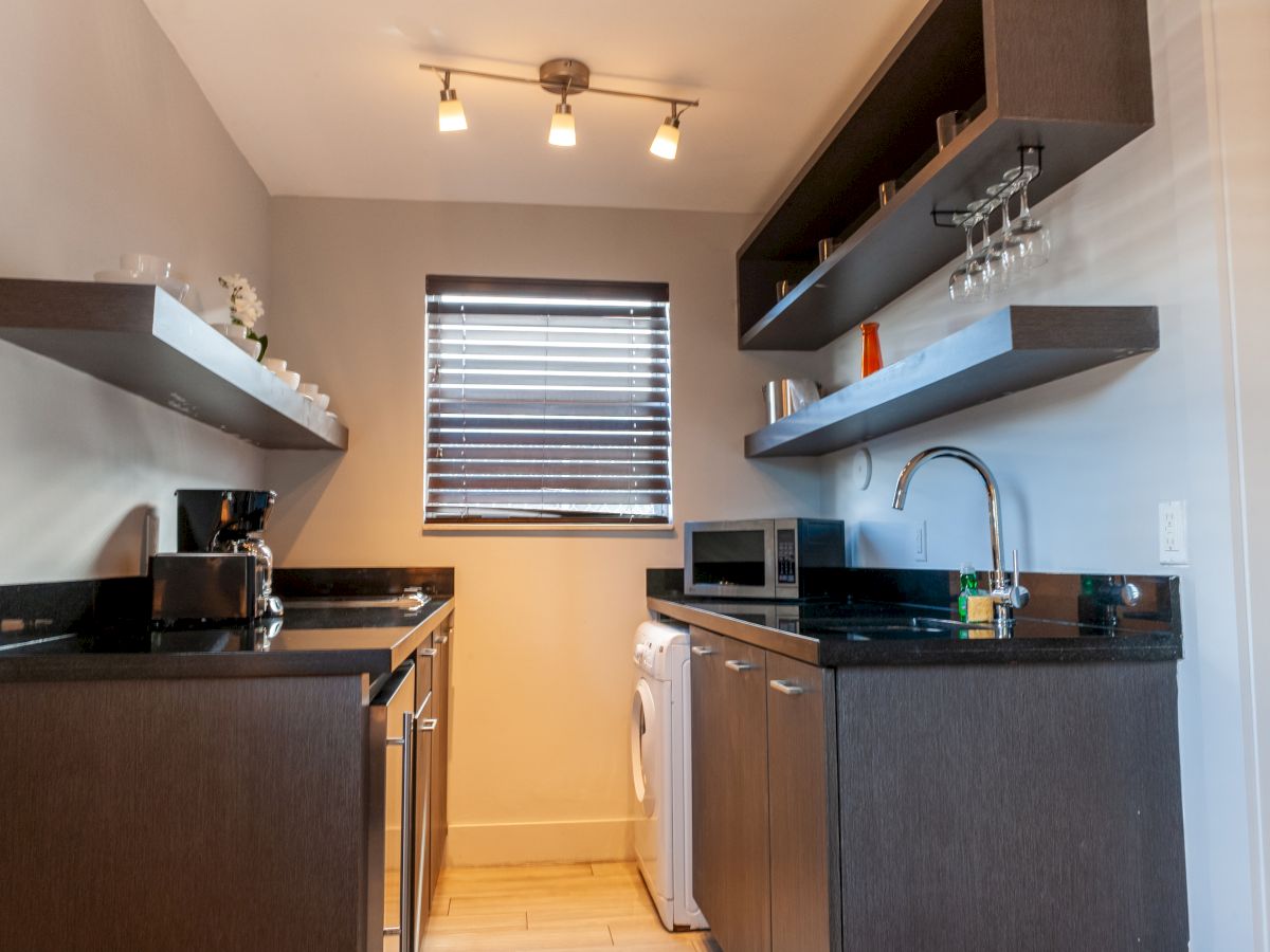 A compact kitchen with dark cabinets, black countertops, open shelves, a small window, and stainless appliances including a microwave and dishwasher.