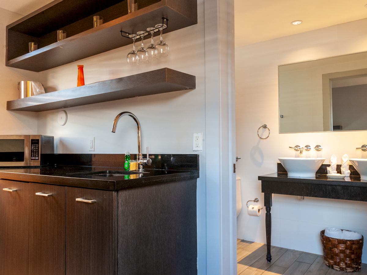 A modern kitchenette with dark cabinets, a black countertop, a sink, and open shelves; a separate vanity area with mirrors and sinks across.