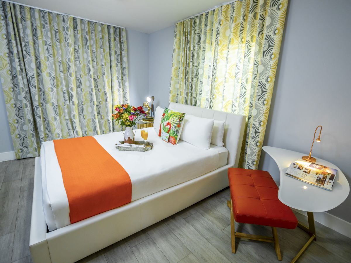 A bright hotel bedroom with a white bed, orange throw, floral curtains, a small desk and red cushioned bench, and a bouquet on the nightstand.