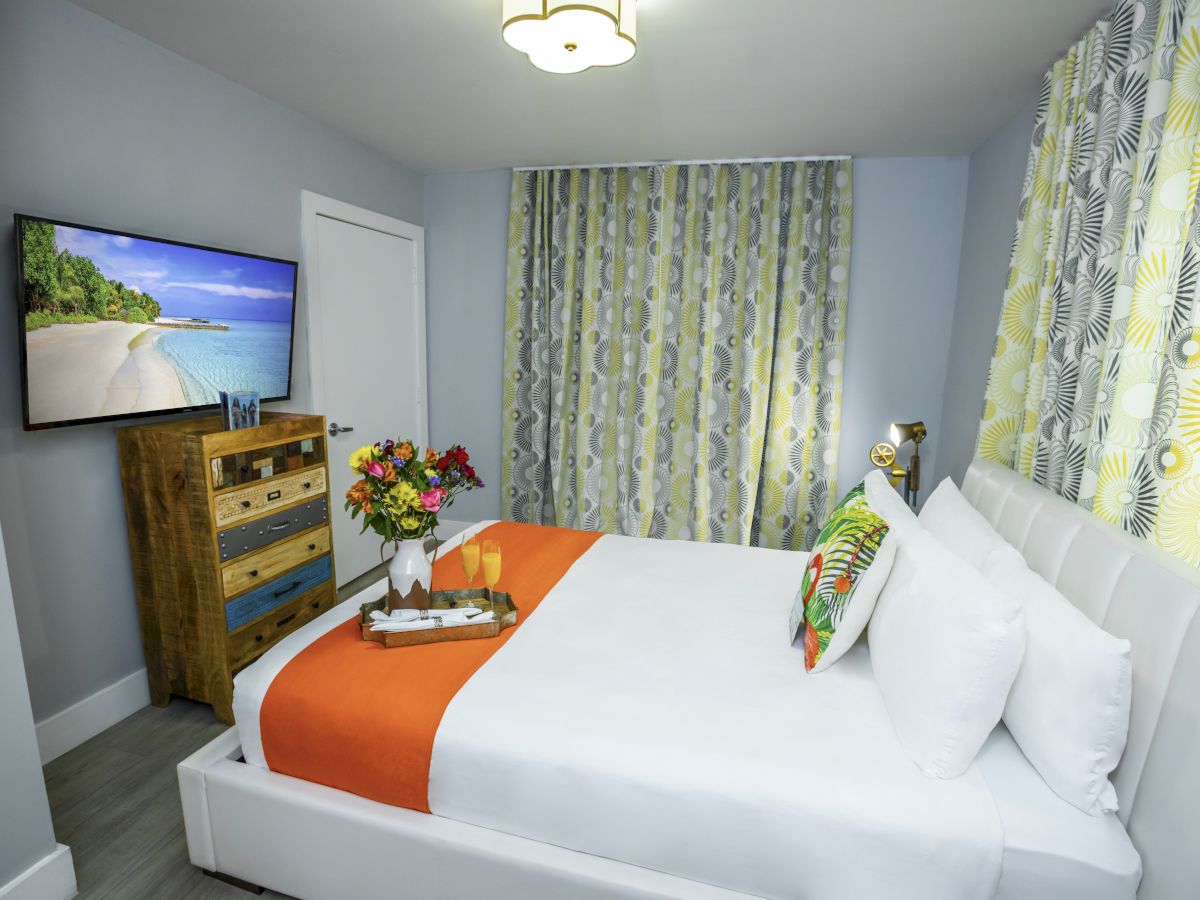 A cozy white bed with an orange runner, colorful pillows, a wooden dresser with a TV, a vase of flowers, and tropical green-yellow curtains.