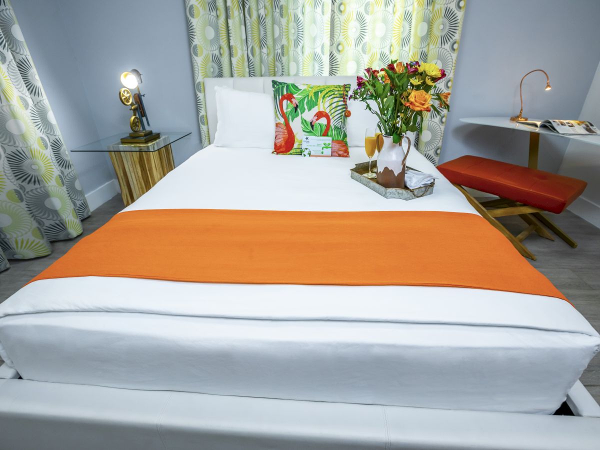 A modern bedroom with a white bed, orange runner, colorful pillows, a small desk and lamp, a bench, and fresh flowers on the nightstand.