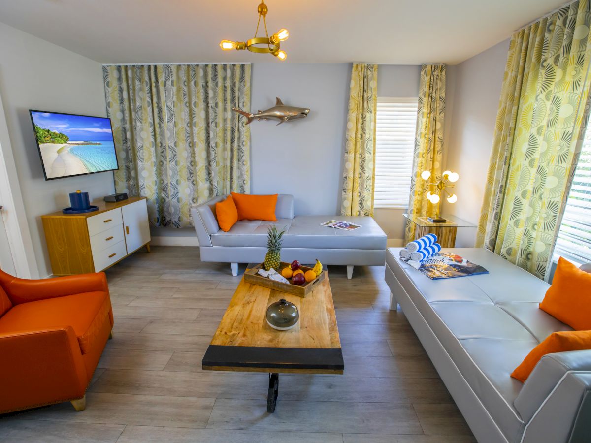 Living room with light gray sectional, orange pillows and chair, wooden coffee table, wall-mounted TV, bright patterned curtains, coastal decor.