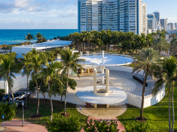 A coastal urban area with modern buildings, a circular structure, palm trees, and a blue ocean backdrop under a partly cloudy sky.