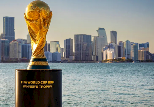 A FIFA World Cup Winners&rsquo; Trophy sits on a pedestal by the water with a city skyline in the background.