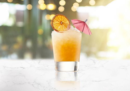 A frosty tropical cocktail with orange gradient, a citrus wheel, and a tiny pink umbrella on a bright, glossy counter.