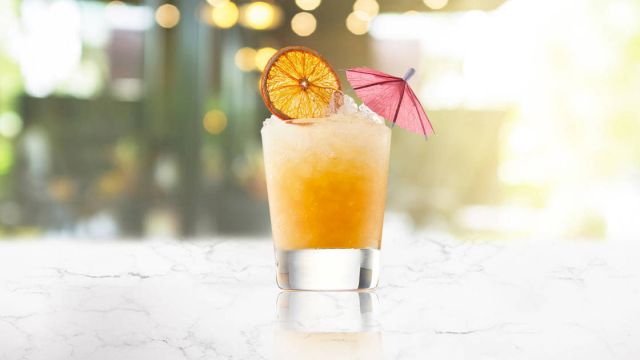 A frosty tropical cocktail with orange gradient, a citrus wheel, and a tiny pink umbrella on a bright, glossy counter.