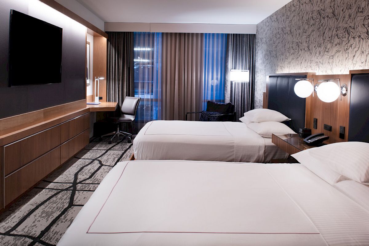 A modern hotel room with two beds, a flat-screen TV, desk, chair, and patterned wallpaper, creating a sleek and inviting atmosphere.