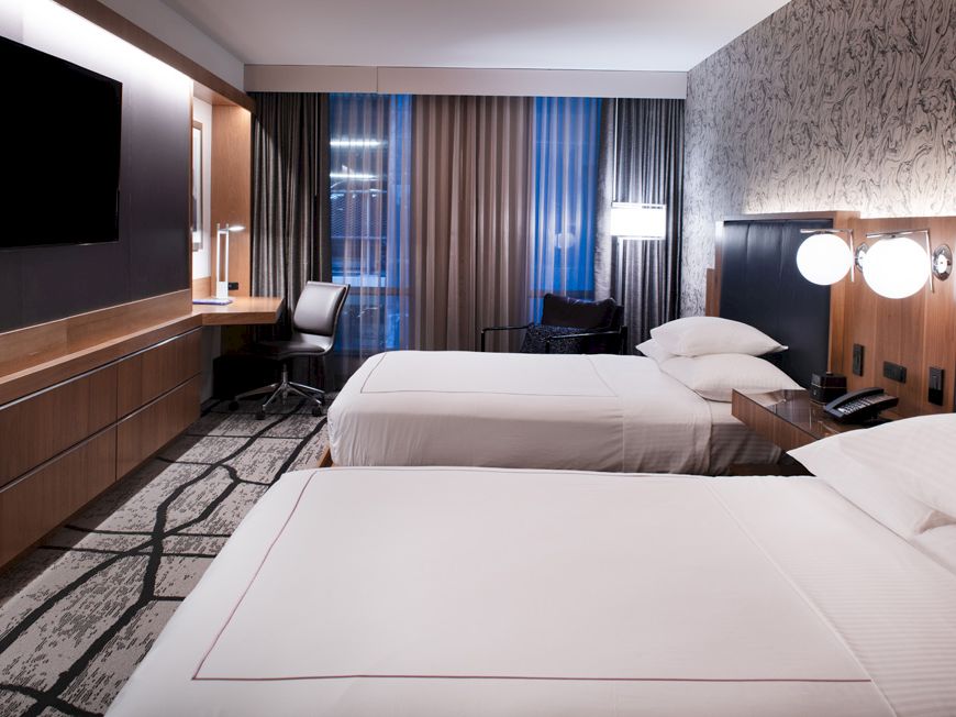 A modern hotel room with two beds, a flat-screen TV, desk, chair, and patterned wallpaper, creating a sleek and inviting atmosphere.