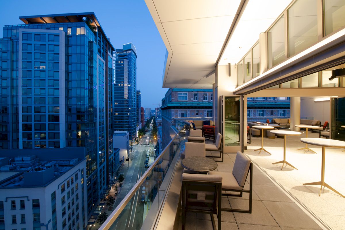 A modern cityscape at dusk with tall buildings and an outdoor balcony featuring seating and tables.