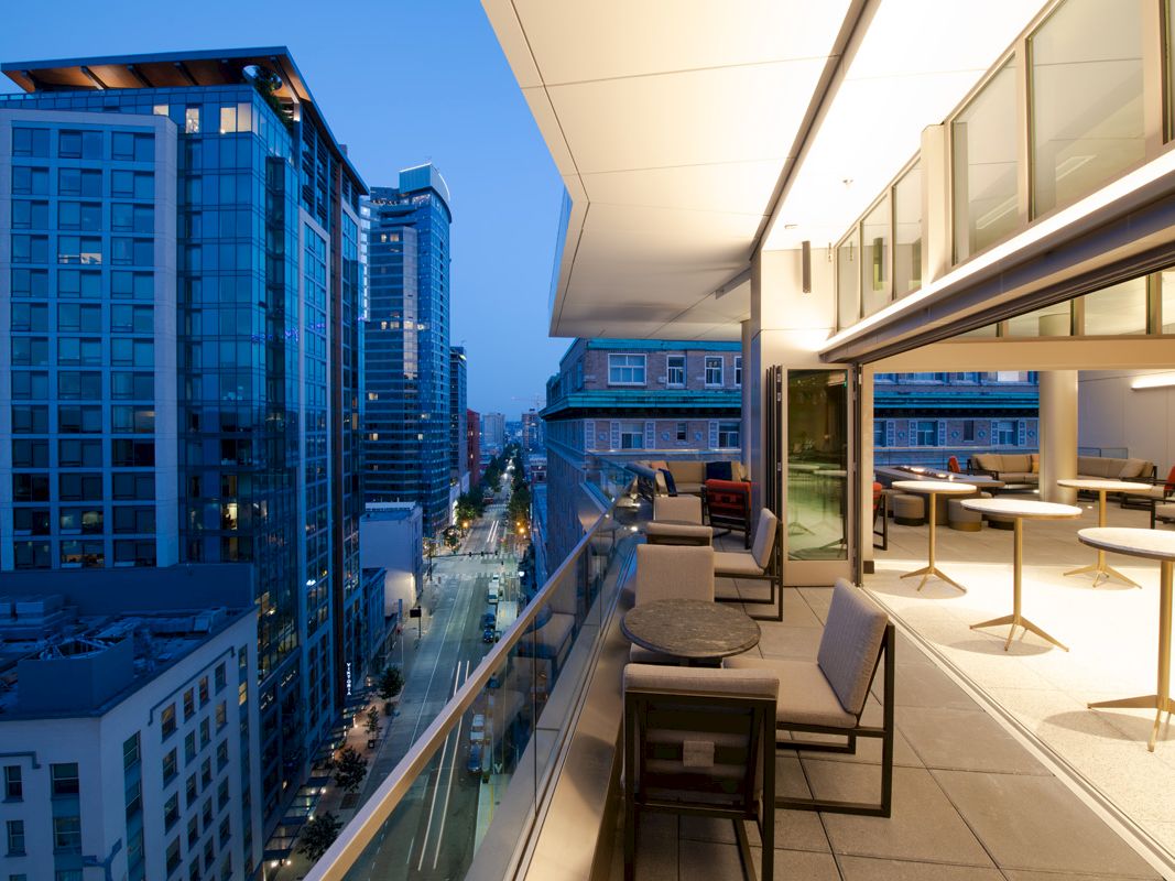 A modern cityscape at dusk with tall buildings and an outdoor balcony featuring seating and tables.