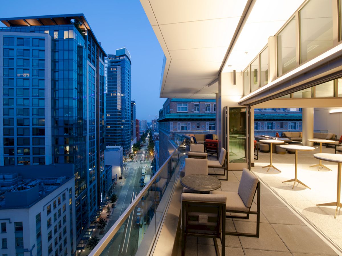 A modern balcony view with tables and chairs, overlooking a cityscape of tall buildings at twilight, creating a serene urban atmosphere.