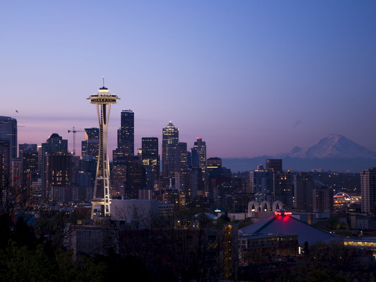 Seattle skyline at dusk with the Space Needle prominently featured and Mount Rainier in the background against a clear twilight sky.
