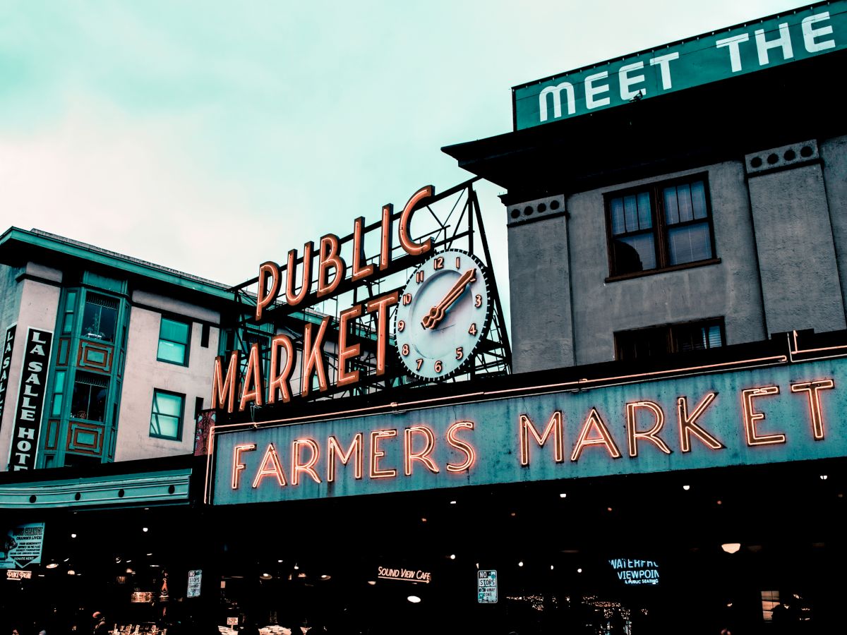 The image shows a famous public market with neon signs that read "Public Market" and "Farmers Market," along with a large clock above.