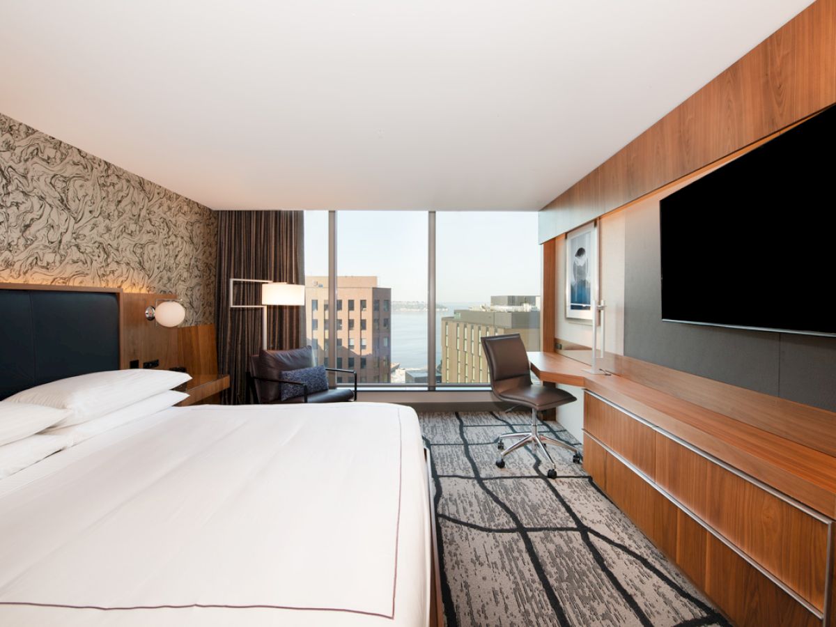 A modern hotel room features a large bed, desk with chair, and a big TV, with a window view of city buildings and water.