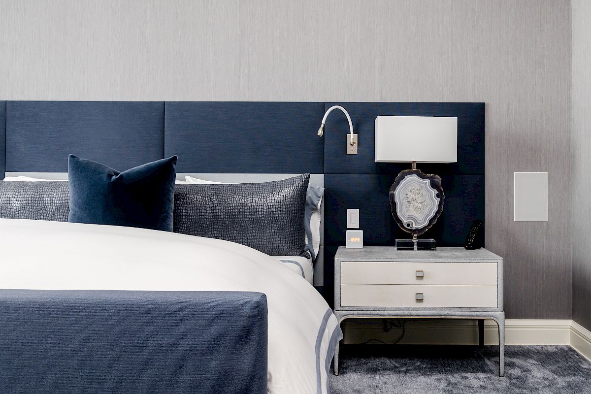 A modern bedroom with a blue headboard, white bedding, pillows, and a nightstand featuring a lamp and decorative object on top.