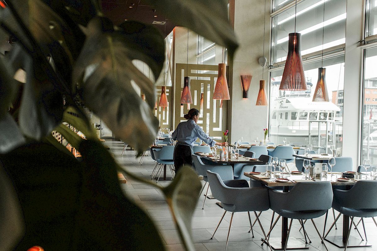 The image shows a modern restaurant interior with a person setting tables, surrounded by plants and stylish lighting.