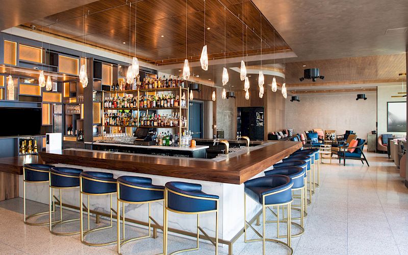 A modern bar with a central counter, stylish lighting, bottles on shelves, and blue cushioned stools, set in an elegant lounge area.