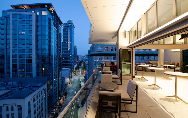 A modern rooftop terrace with seating overlooks a cityscape of tall buildings and a street, lit up during twilight.